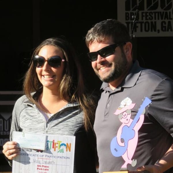 Two smiling people in sunglasses holding a Holy Smokes BBQ award for 5th Place Pork Loin.