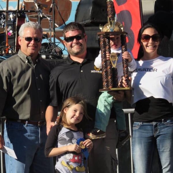 Two men, two women, and two children pose with a large trophy and a blue ribbon.