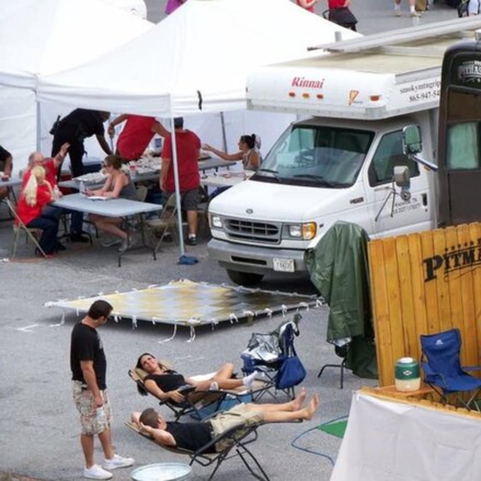 People relaxing in chairs and under tents next to a white Holy Smokes BBQ catering truck.
