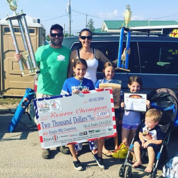 A family of five, including three children, proudly displays trophies and a large check for $2,000.
