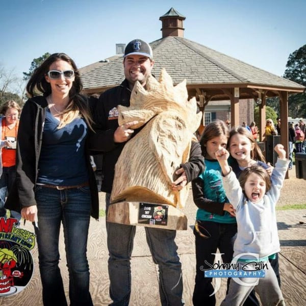 A family poses with a large wooden trophy, celebrating a Holy Smokes BBQ win.