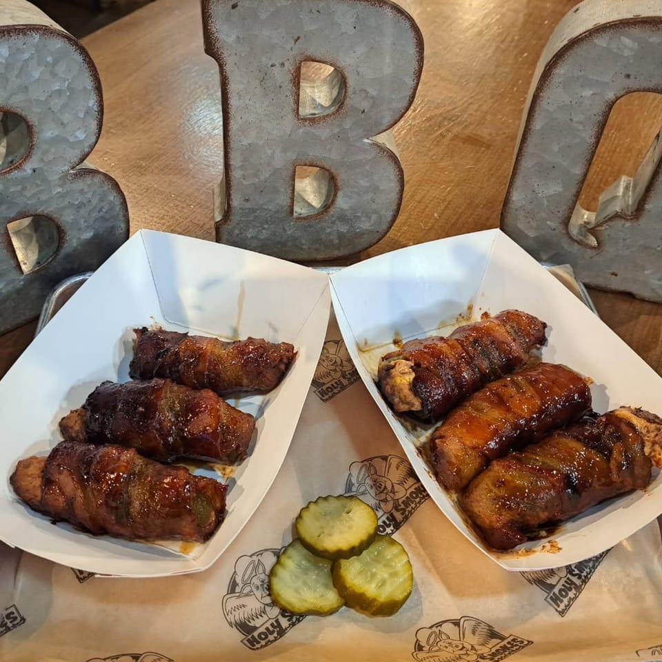 Two trays of Holy Smokes BBQ bacon-wrapped chicken, with pickles and large metal "BBQ" letters.