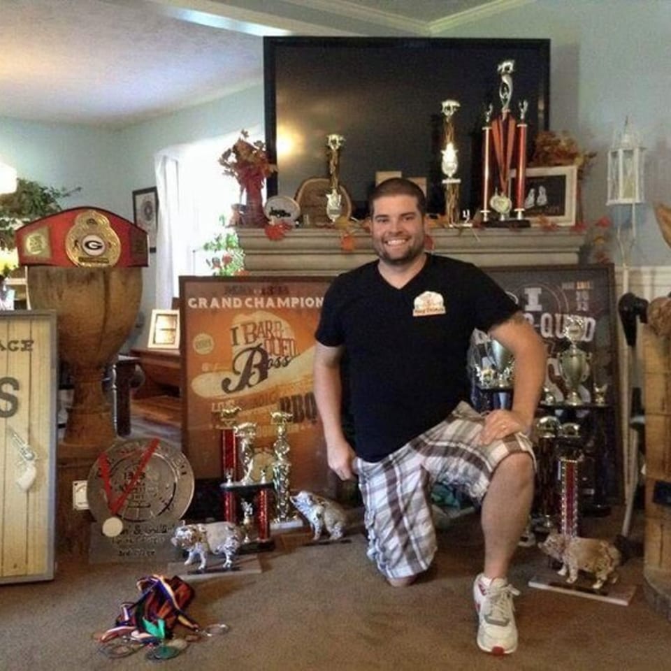 A man kneels among numerous BBQ competition trophies, including a championship belt and medals.