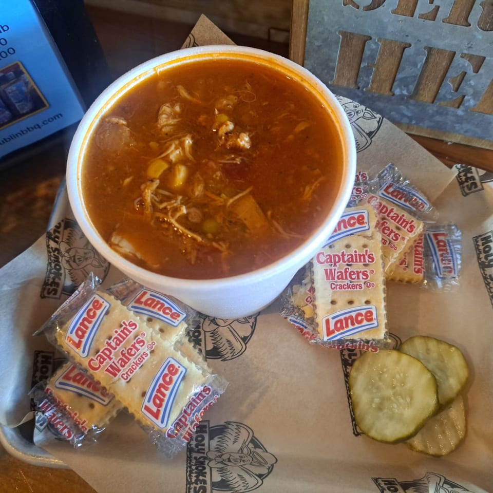 A bowl of Holy Smokes BBQ Brunswick stew with crackers and pickles on a tray.