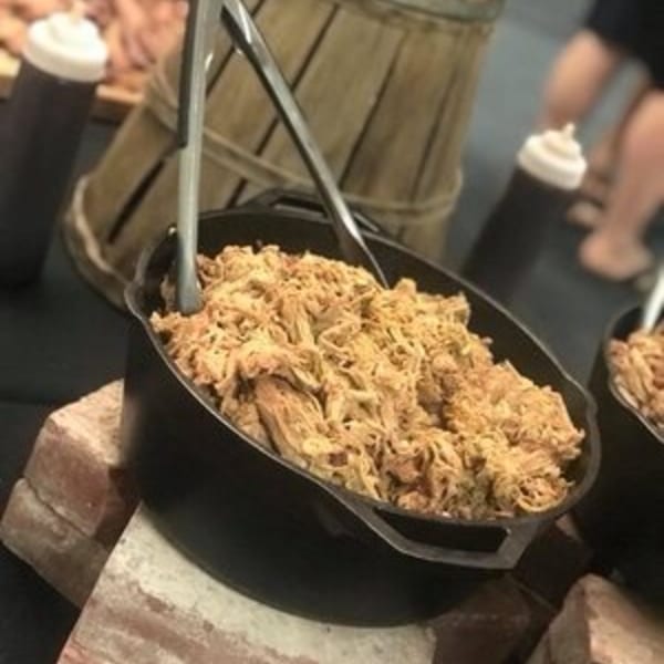 A cast iron pot filled with Holy Smokes BBQ pulled pork, with tongs and sauce bottles.