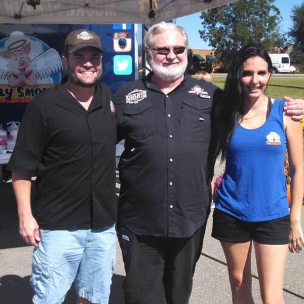 Three Holy Smokes BBQ team members, two men and one woman, smiling outdoors.