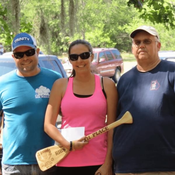 Three Holy Smokes BBQ team members, two men and one woman, pose outdoors with a "Ribs 2017" award.