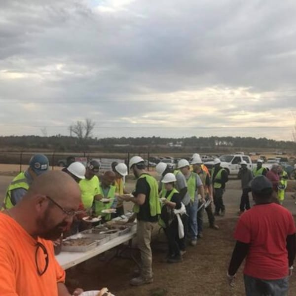 Construction workers in hard hats and safety vests line up for Holy Smokes BBQ at an outdoor event.