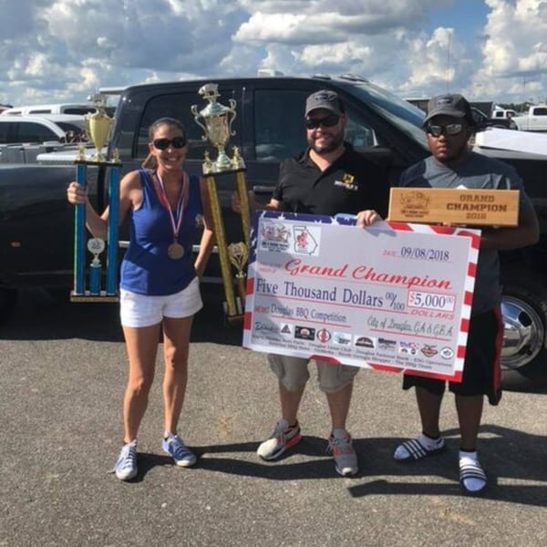Three people holding trophies, a large check for $5,000, and a "Grand Champion" plaque from a BBQ competition.