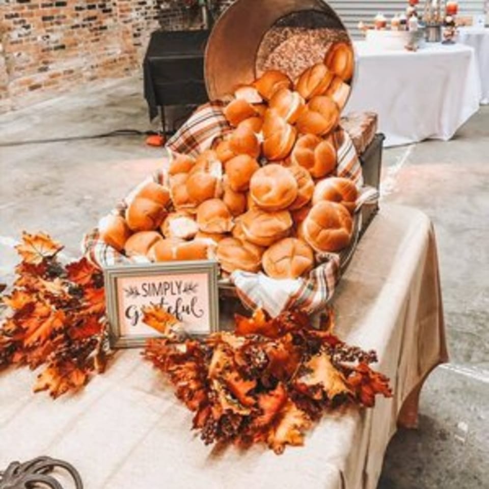 A large basket overflowing with dinner rolls, decorated with autumn leaves and a "Simply Grateful" sign.