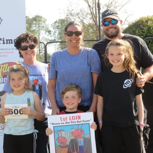 A family of five, including three children, smiling and holding awards outdoors at a Holy Smokes BBQ event.