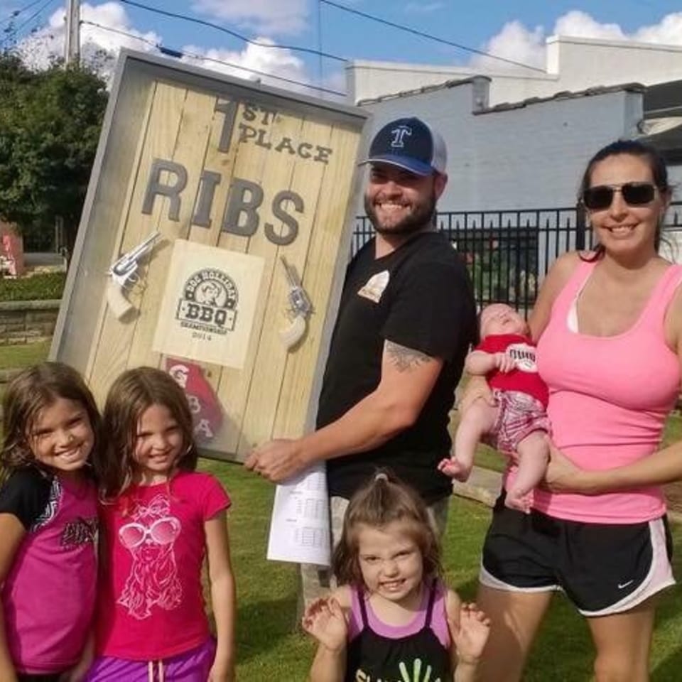 A family of six, including a baby, poses with a large "1st Place Ribs" sign outdoors.